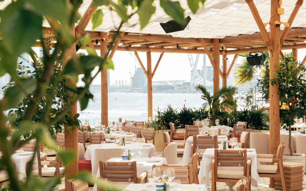Wide-angle view of the 363 m² Cape Reed timber pergola at Giulia by the Beach restaurant, Bebé Beach, Dubai Harbour — showing the full span of cylindrical columns, lath canopy, canvas shade sails, and lush coastal planting against the Dubai waterfront