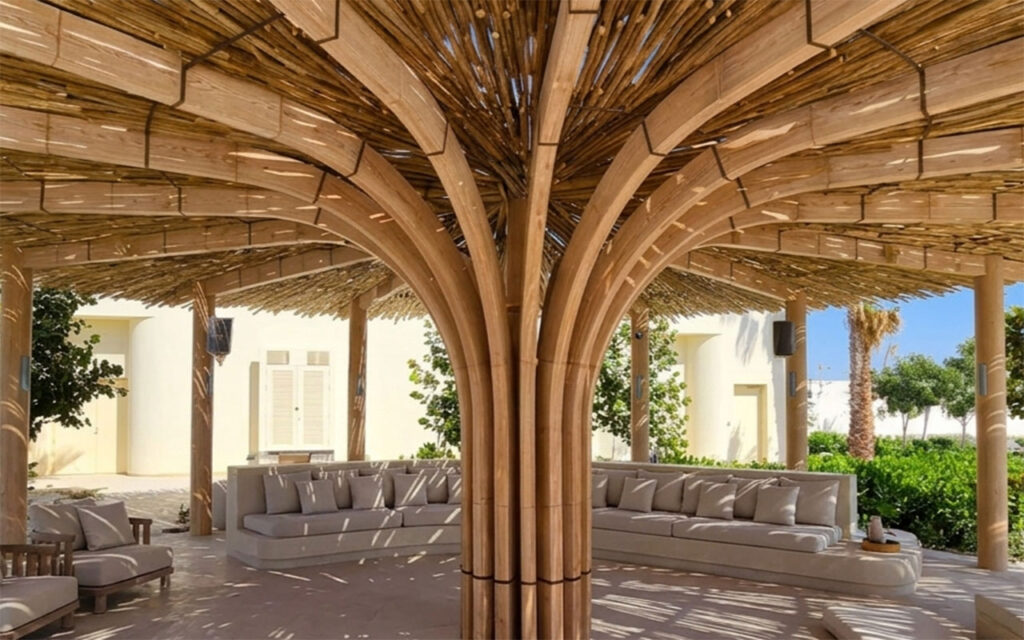 Cape Reed timber-lath kiosk at Shura Island resort, Saudi Arabia, showing arched wooden columns supporting a woven lath canopy roof with dappled sunlight and neutral outdoor seating below